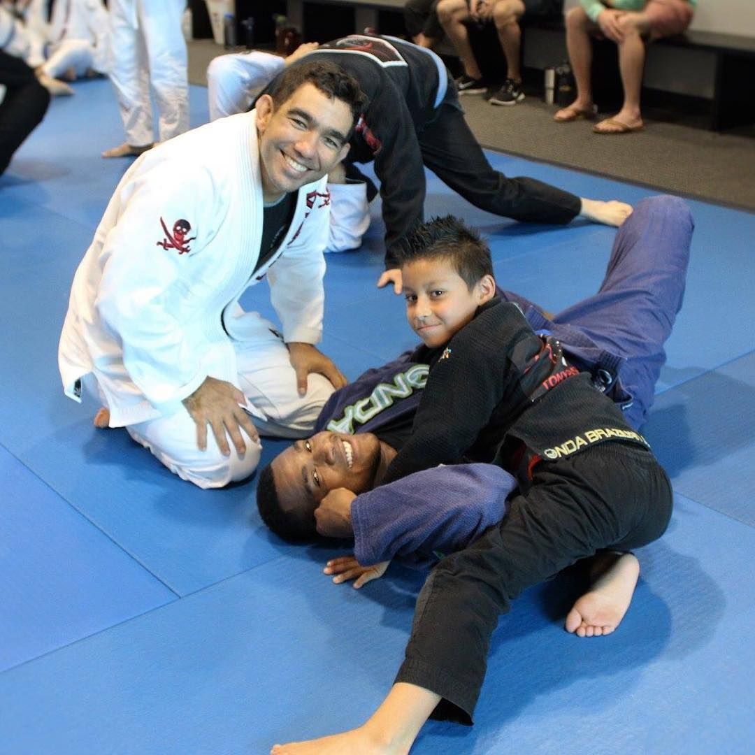 A group of people in a jiu-jitsu class. An instructor kneels, smiling at the camera, watching a student.
