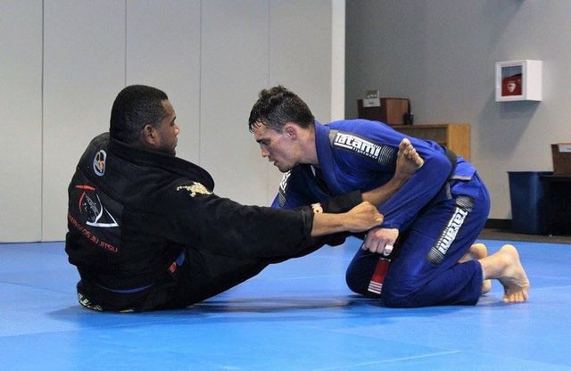 Two men in martial arts gear grappling on a blue mat, one with a leg lock attempt.