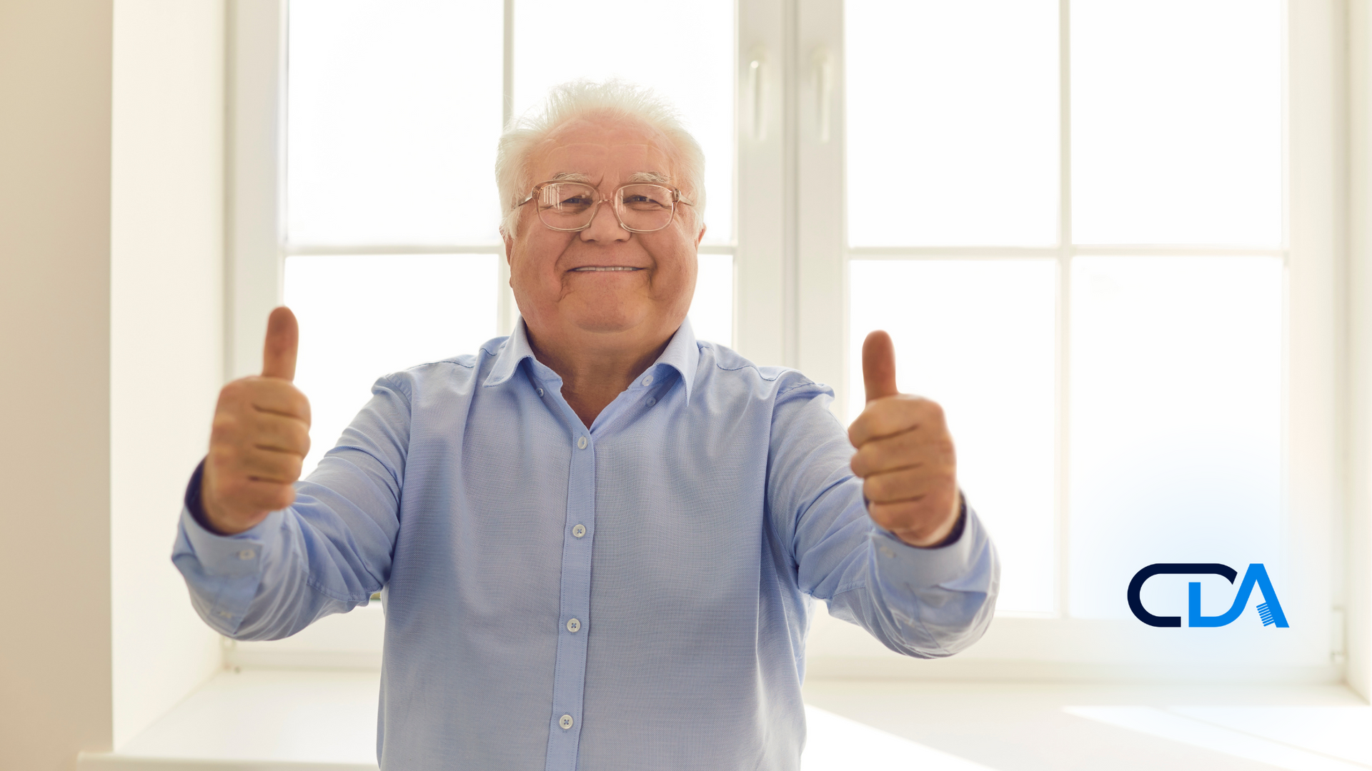 An elderly man is giving two thumbs up in front of a window.