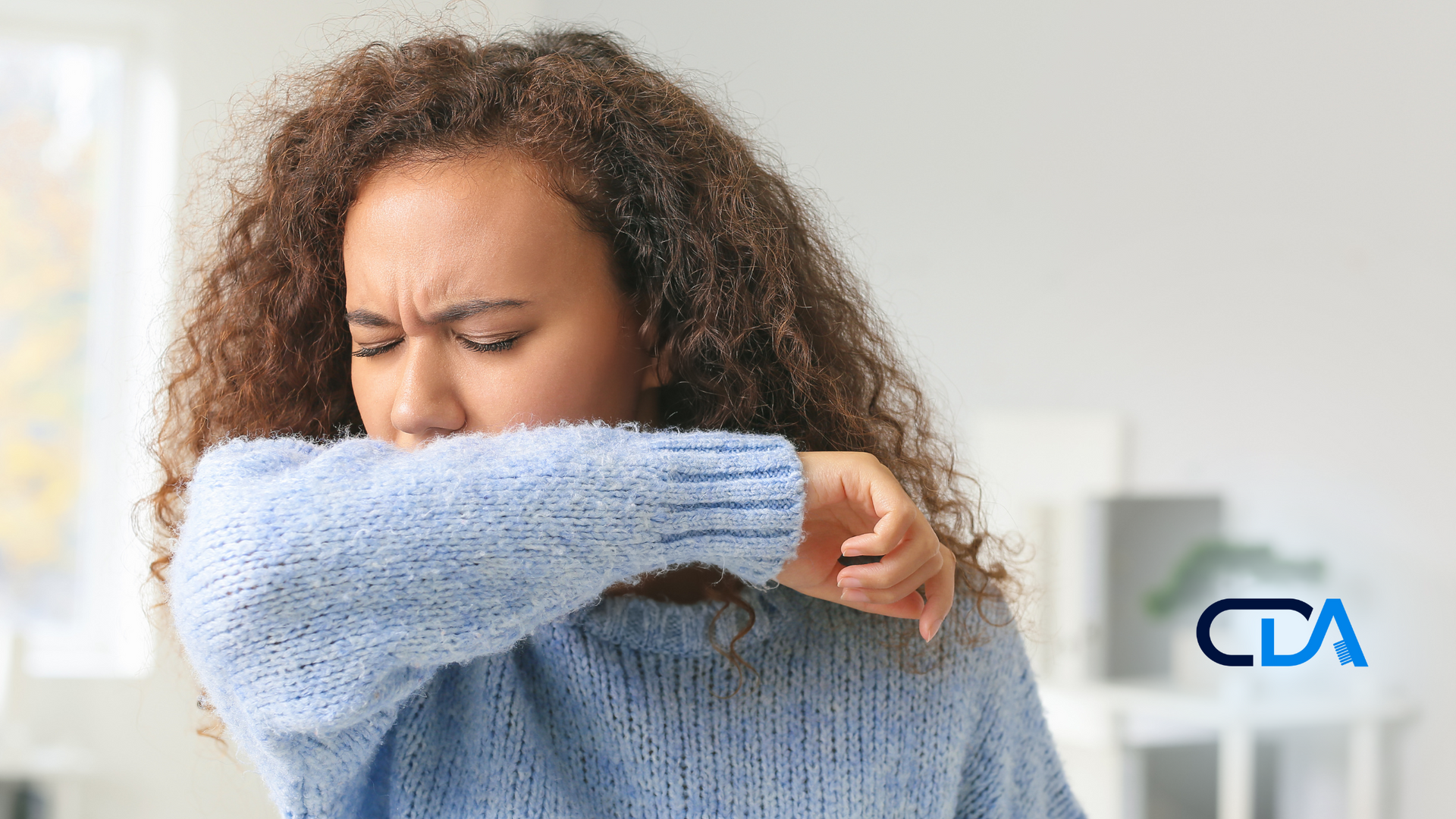 A woman is coughing into her elbow while wearing a blue sweater.