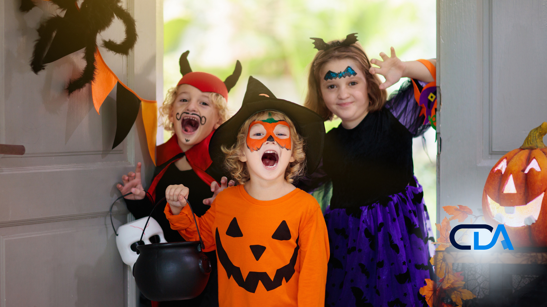 Three children in Halloween costumes excitedly stand at a doorway with decorations, orange pumpkin-themed attire, and cauldron.