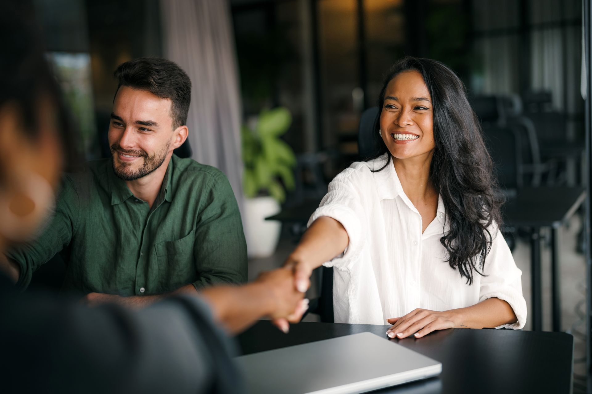 A person shakes hands with another in an office setting, while a third individual looks on and smiles.