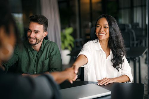 A person shakes hands with another in an office setting, while a third individual looks on and smiles.