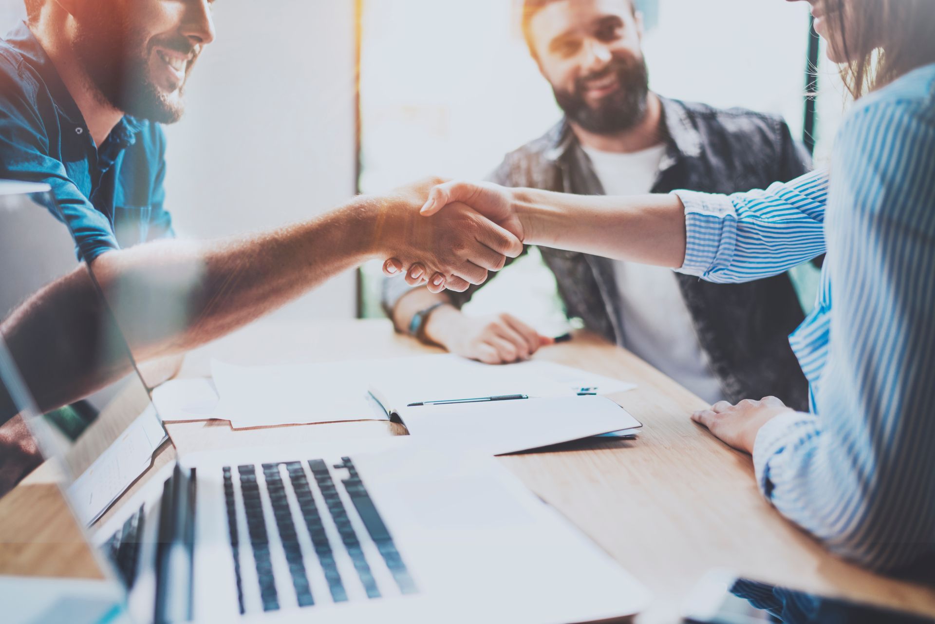 Two people shake hands over a desk with a laptop and documents, while a third person looks on in a bright office.