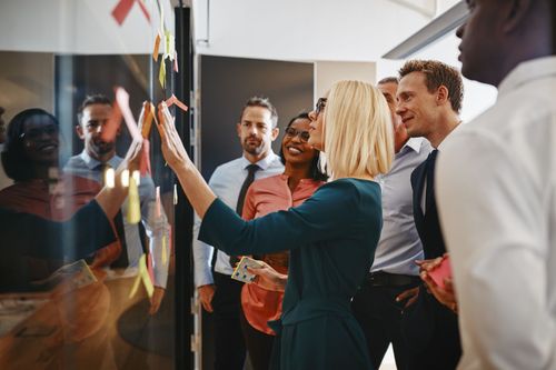 A group of colleagues collaborates at a glass office partition, posting colorful sticky notes during a brainstorming session.