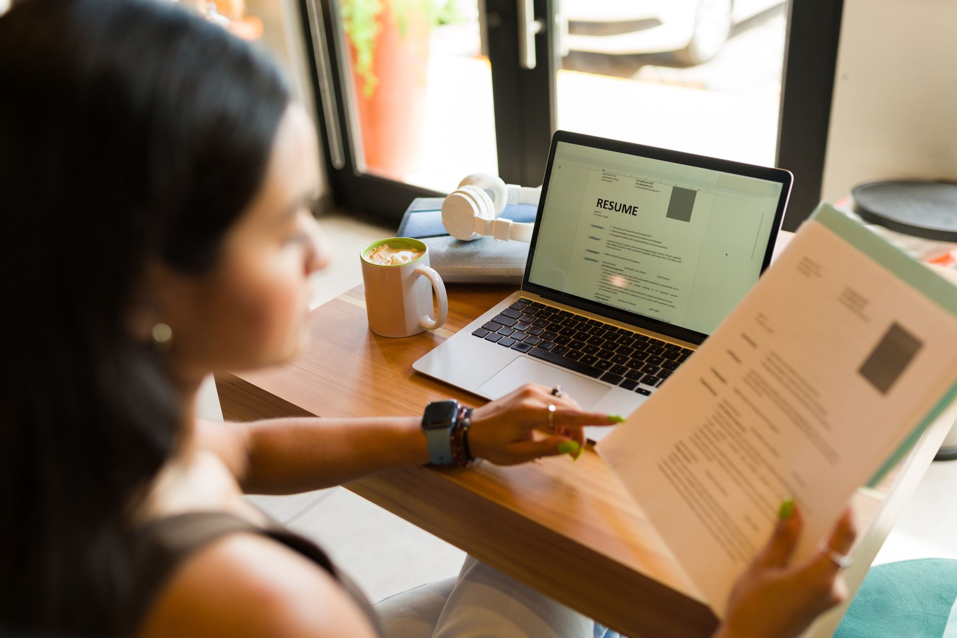 Person reviewing a document beside a laptop at a sunlit table, with a mug and window in the background