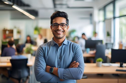 A smiling professional with glasses and folded arms stands in a modern, blurred office setting.