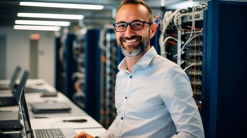 A person with a beard and glasses smiling in a server room, standing near a rack of computer equipment.