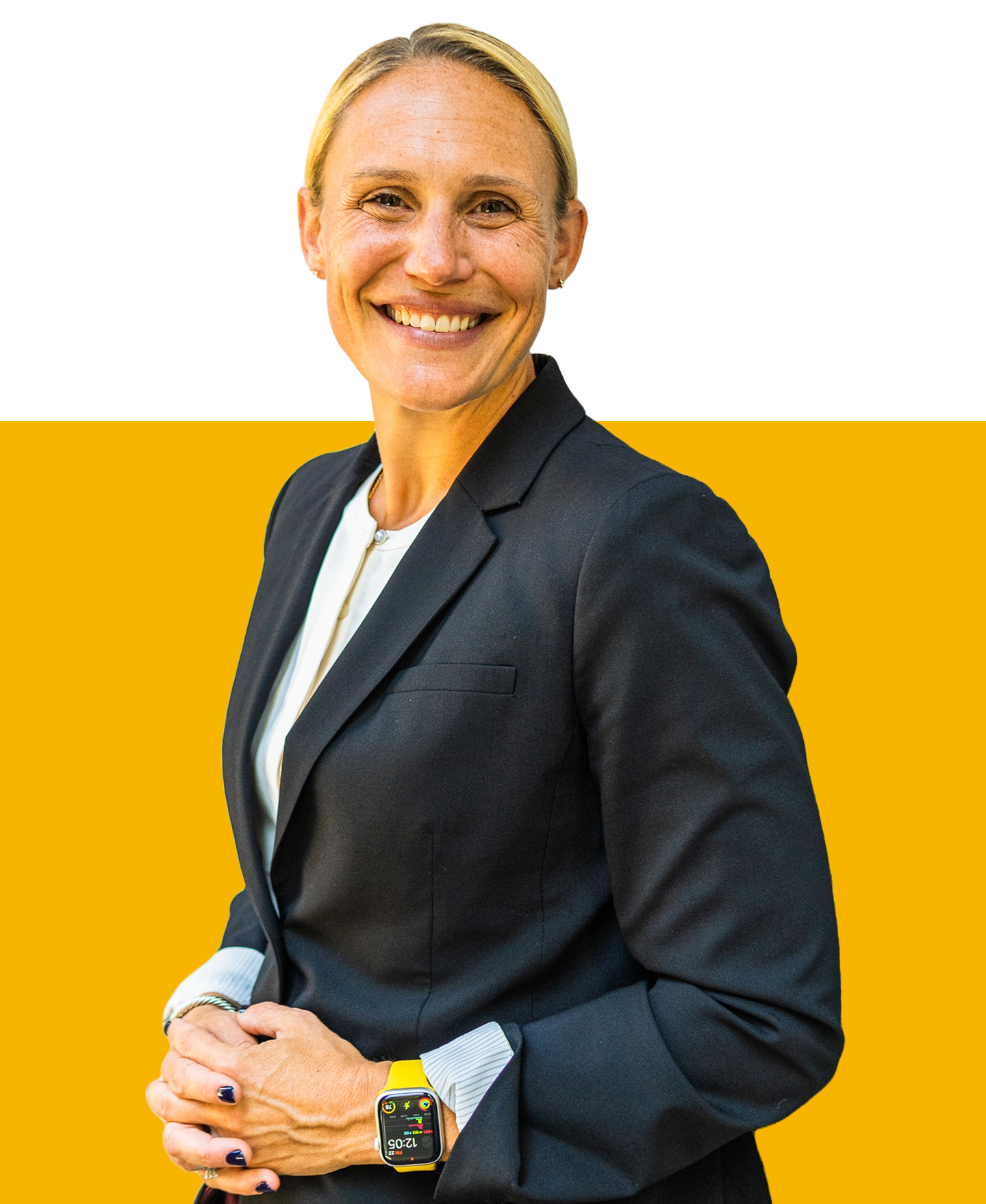 A smiling individual in a dark blazer and white shirt stands before a solid yellow background, hands clasped together.
