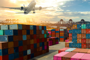 Shipping containers stacked in a port, with a plane taking off overhead against a sunset sky.