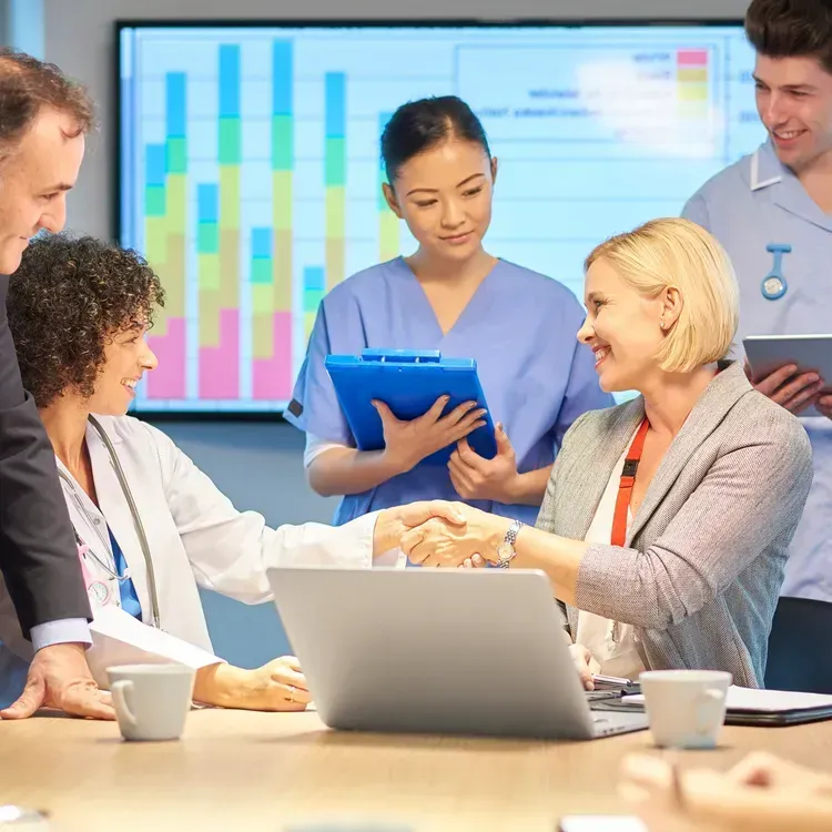 A group of professionals in a meeting room with a chart in the background, two individuals shaking hands over a laptop.