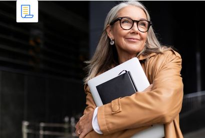 A person with grey hair and glasses in a tan jacket holds a laptop and notebook against their chest, smiling outdoors.