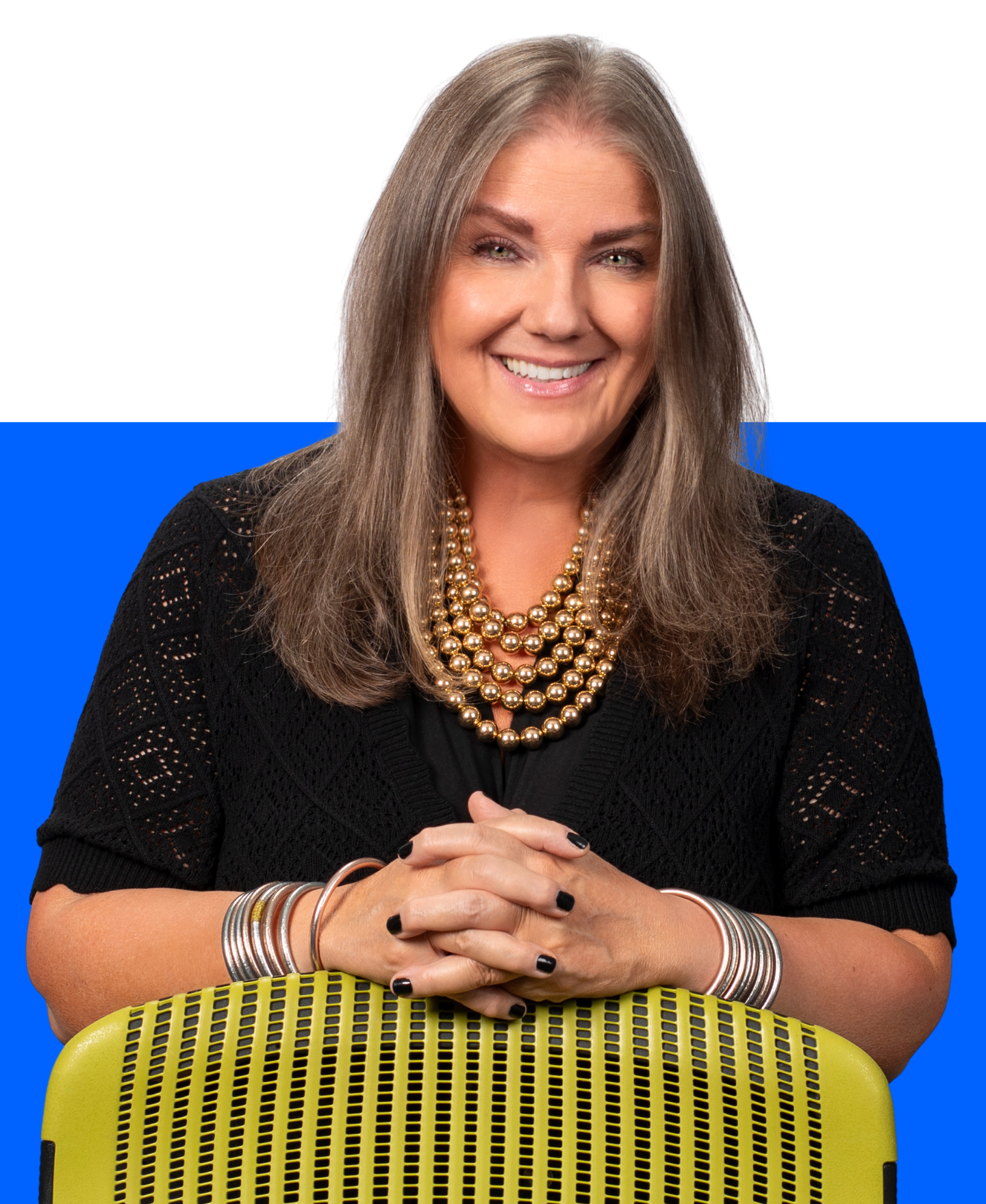 A person with shoulder-length gray hair wearing a dark denim jacket, smiling against a white and blue background.