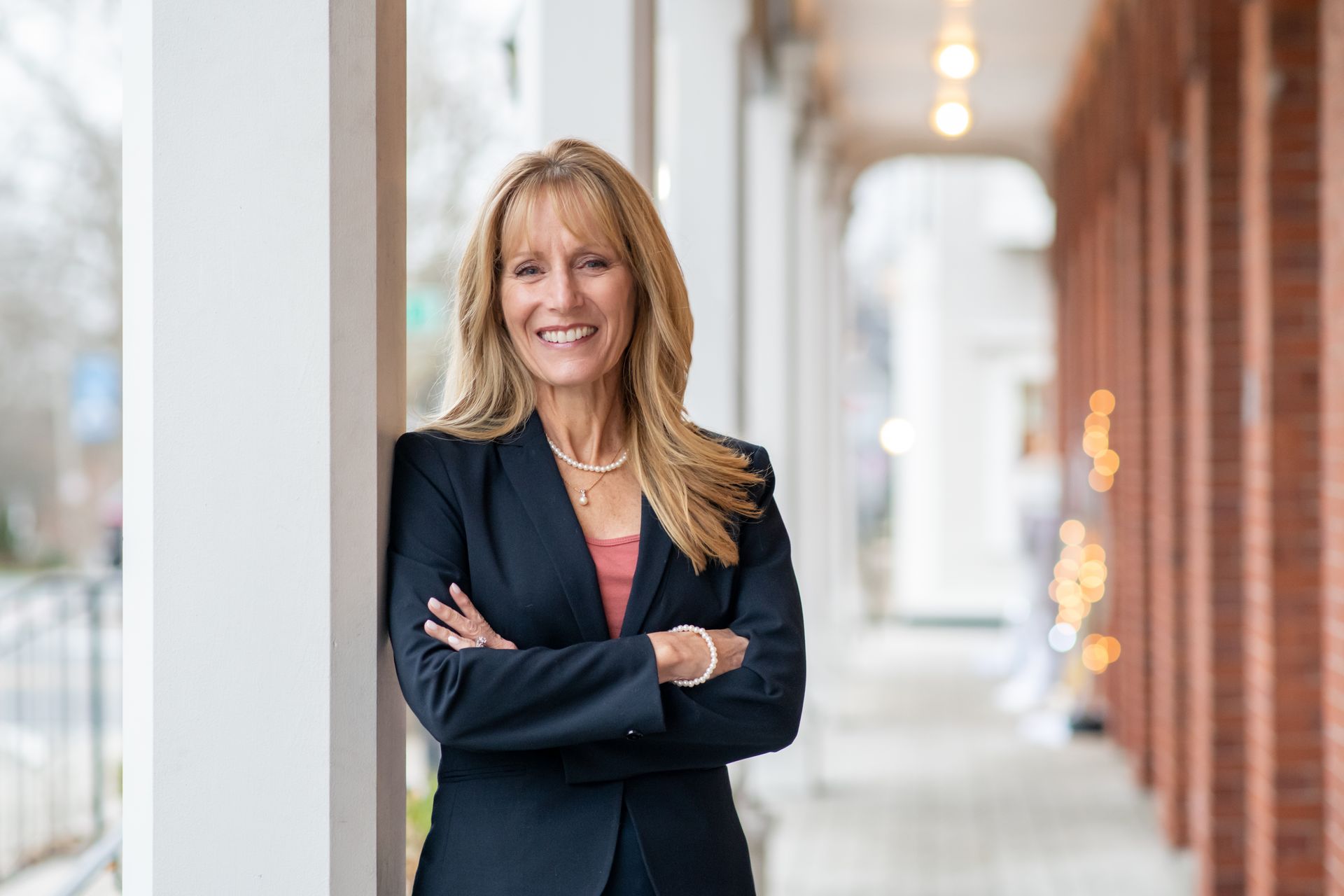 A person with long blonde hair, wearing a dark blazer and necklace, smiles with arms crossed against a solid yellow backdrop.