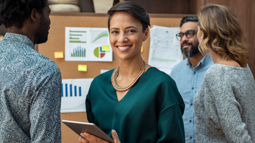 A smiling person holding a tablet, standing in front of a corkboard with charts, surrounded by colleagues in an office.