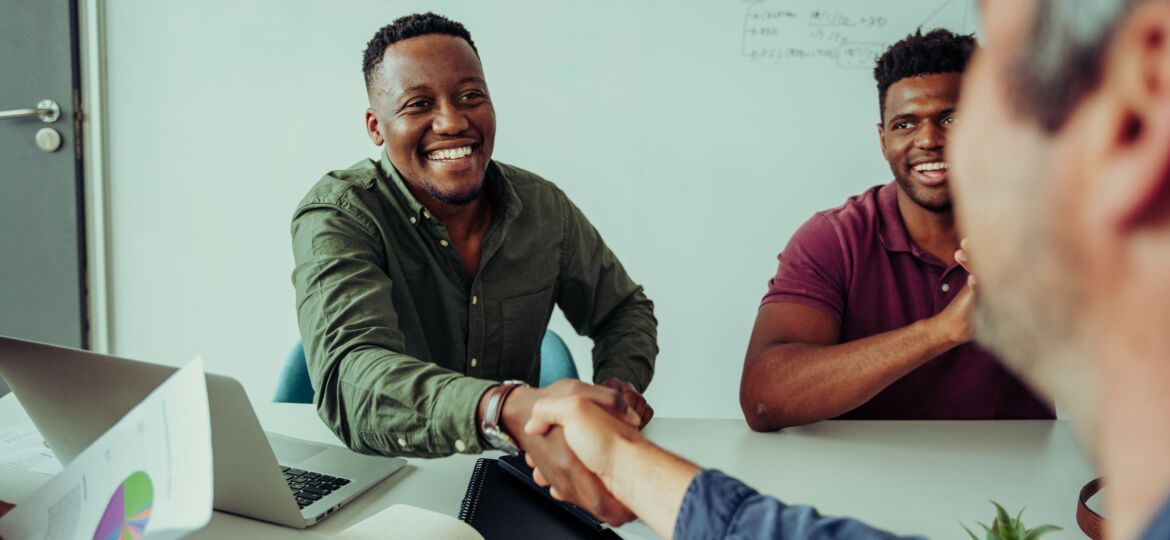 Two colleagues shake hands across an office desk, smiling at a third person, while a laptop and charts sit nearby.