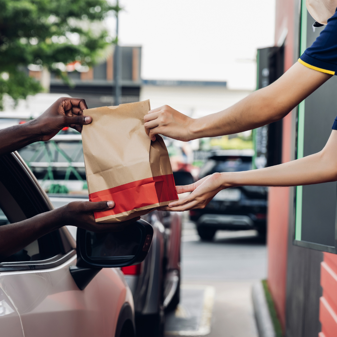 Person in a car receiving a brown paper bag from a fast-food worker at a drive-through window.