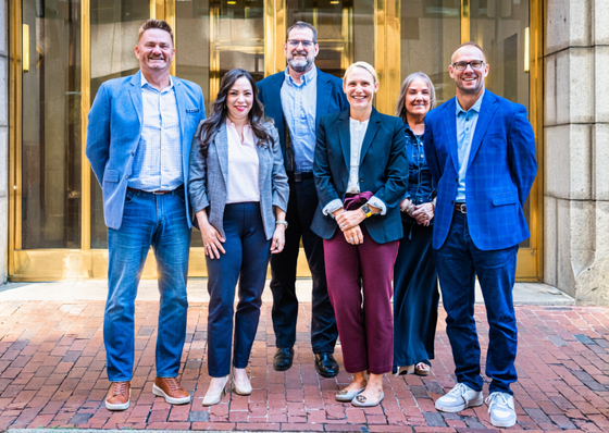 A group of eight people stand together in two rows in front of a building entrance with brick flooring.