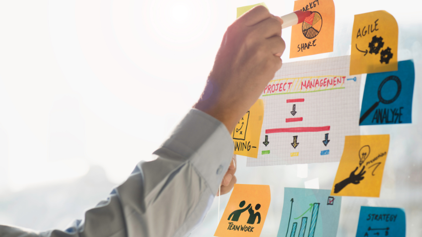 Person in a light shirt writing on a glass pane covered in project management sticky notes.