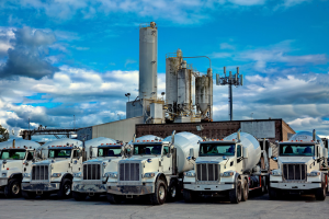 Cement trucks parked in front of a concrete plant under a cloudy blue sky.