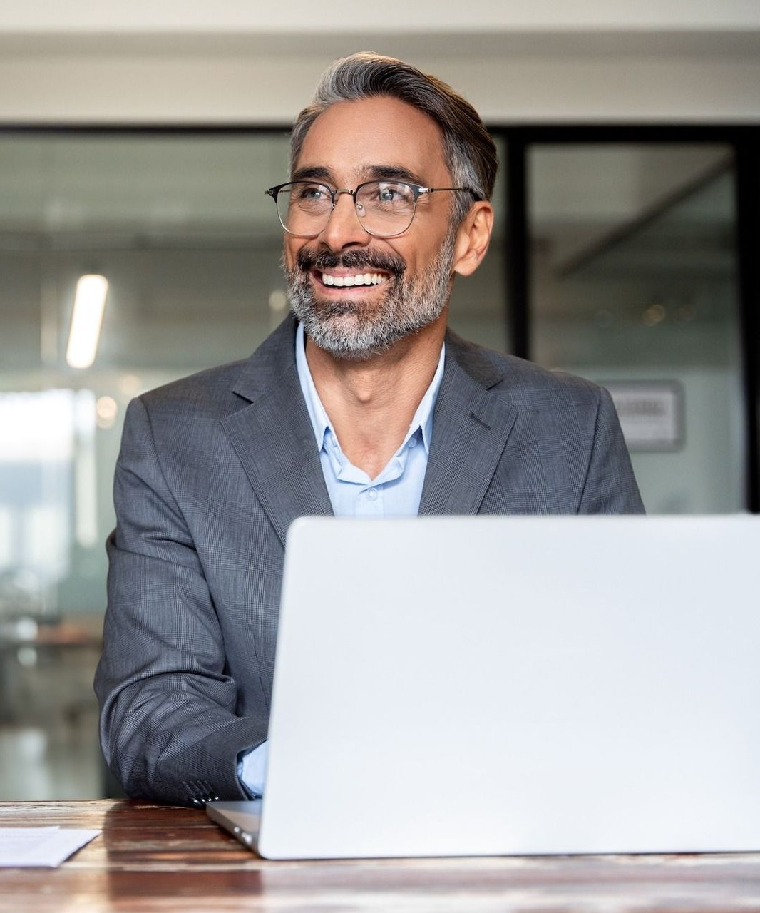 A smiling professional with a beard and glasses sits at a desk in an office, working on a laptop.