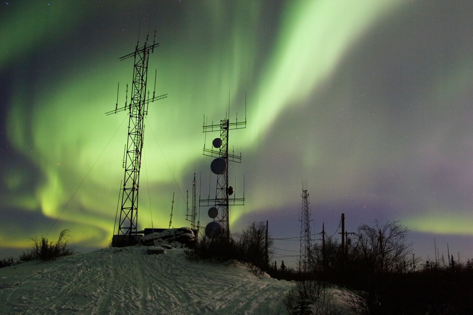Green aurora borealis over communication towers on a snow-covered hill at night in Alaska.