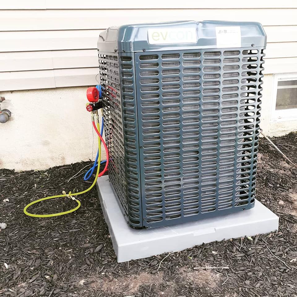 Air conditioning unit outdoors on a gray platform, with gauges and hoses attached; on dark mulch.
