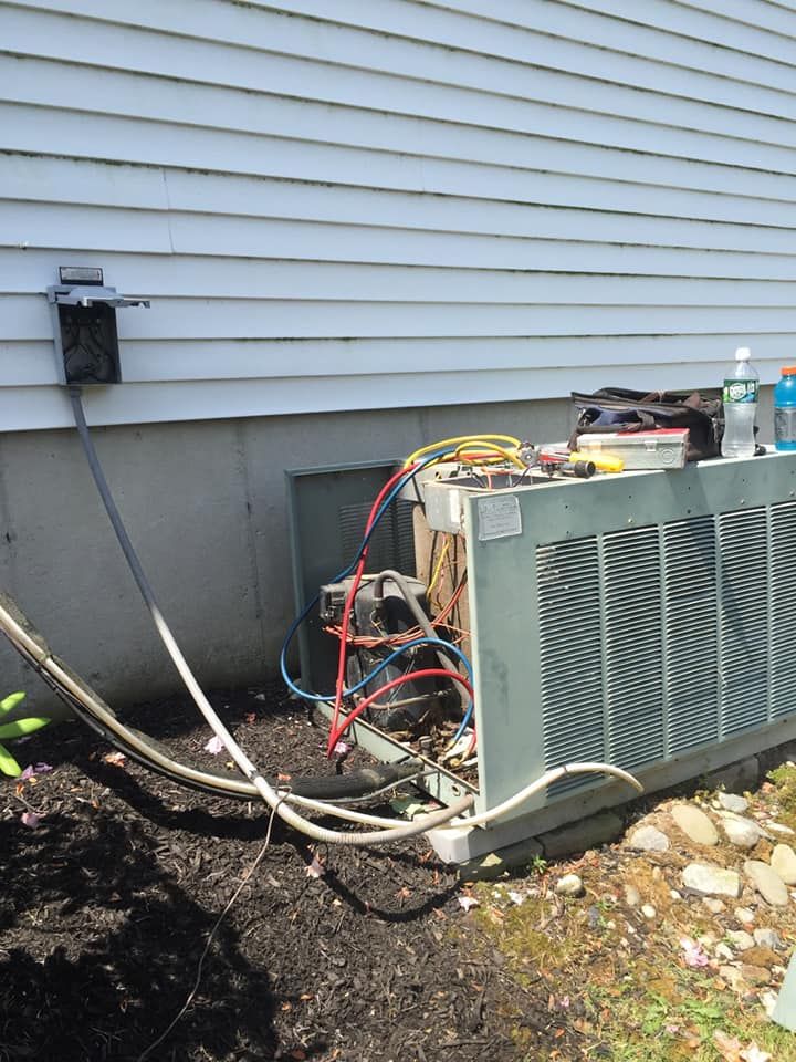 An air conditioning unit being serviced outdoors next to a house. Wires and components are exposed.
