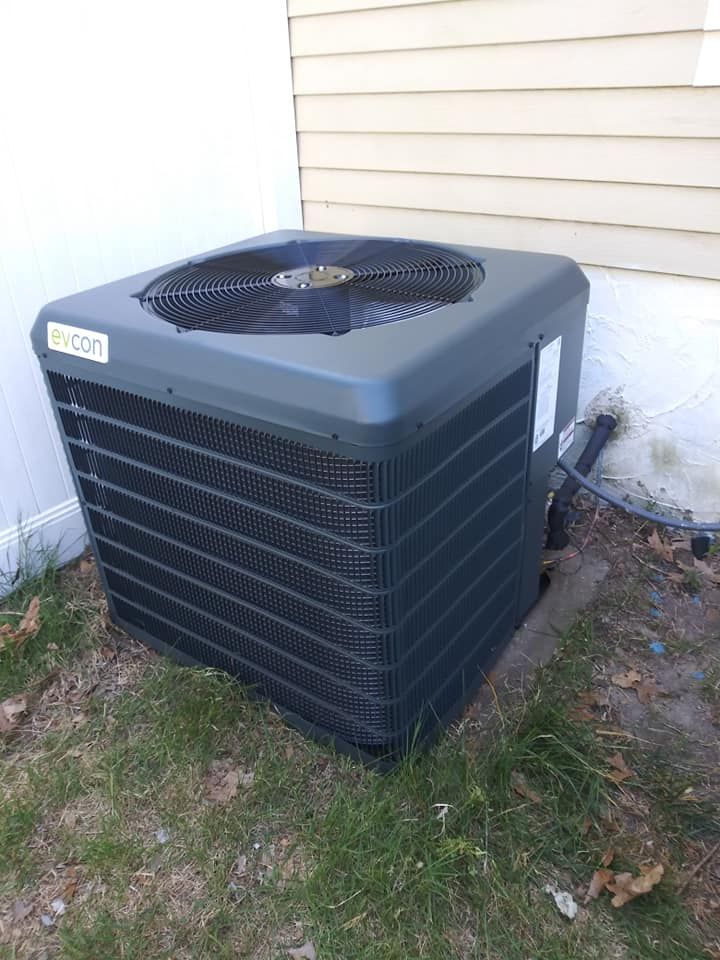 An outdoor air conditioning unit with a fan on top, placed on grass next to a house wall.