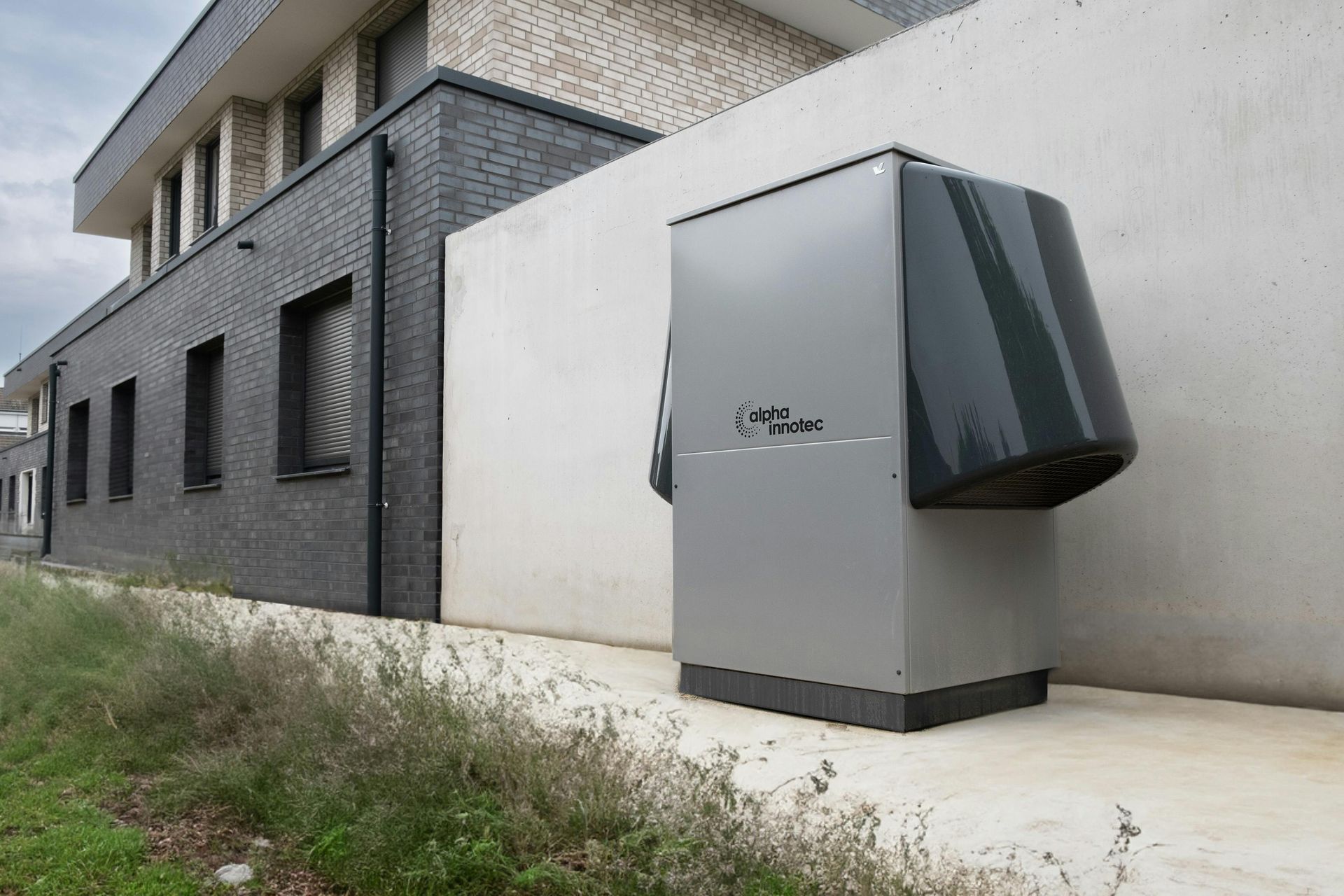Gray heat pump unit next to a light-colored building, with greenery in the foreground.