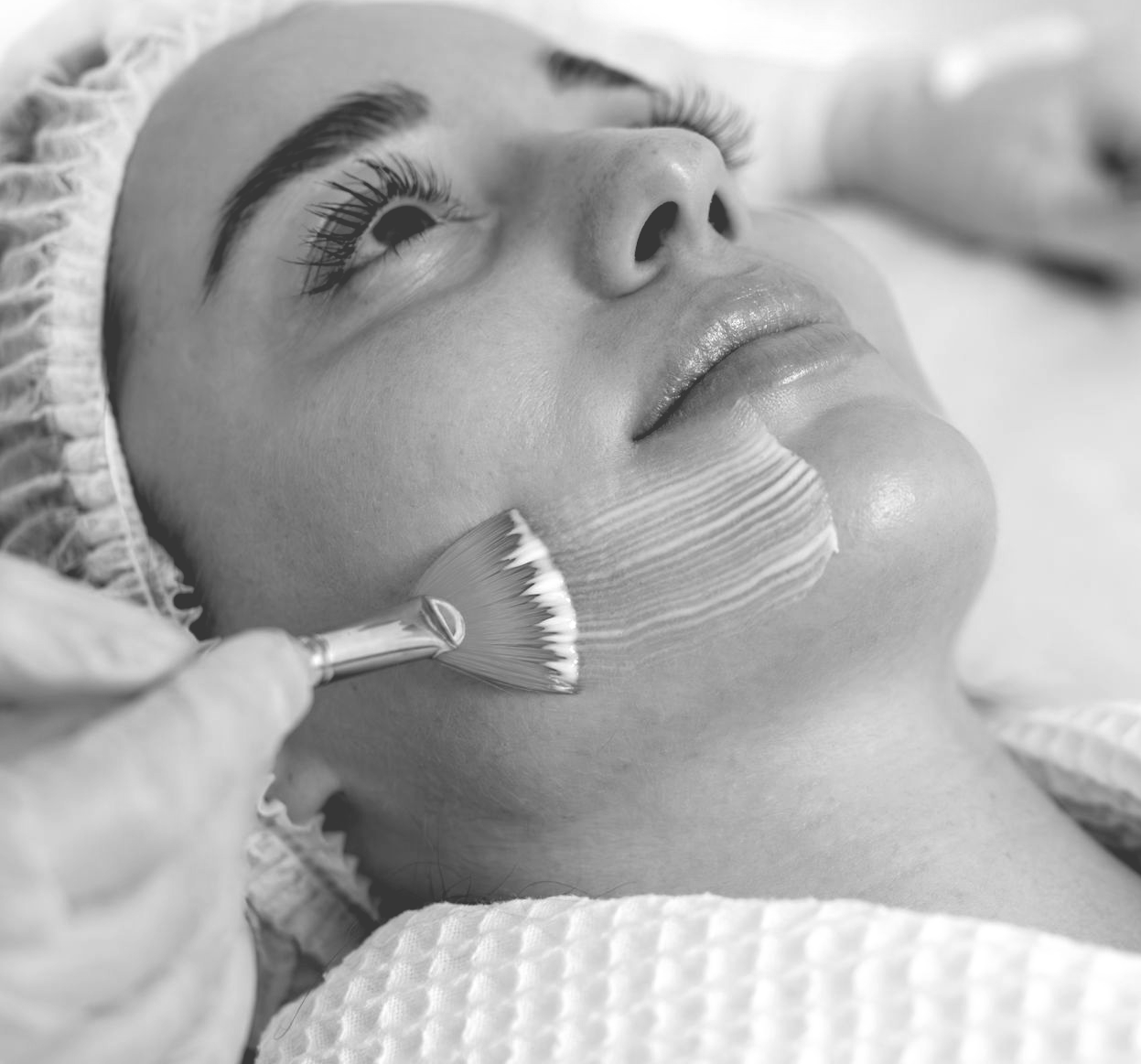 A person receiving a facial peel, with a brush applying a white substance to their cheek.