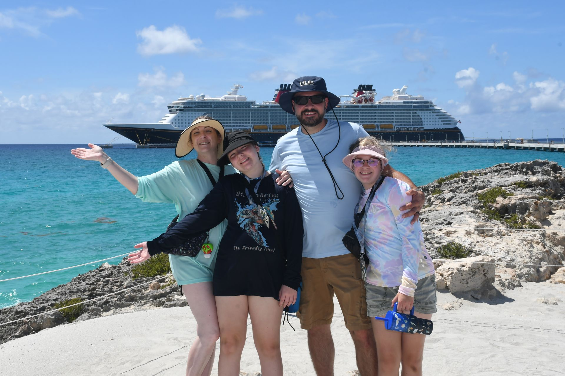 Family poses on a beach in front of a cruise ship; ocean and blue sky in background.