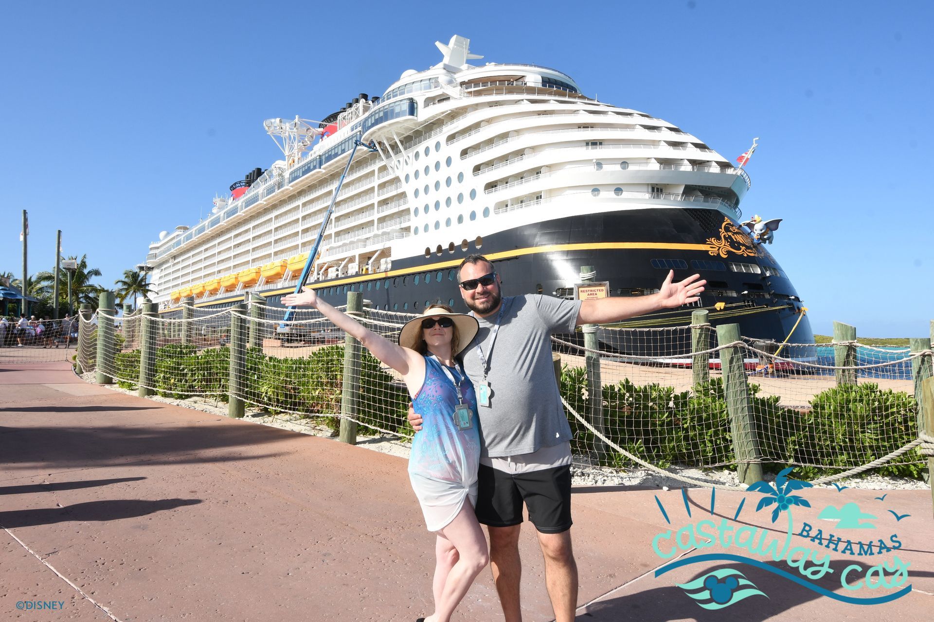 Couple posing in front of a Disney cruise ship with arms outstretched, sunny outdoor setting.