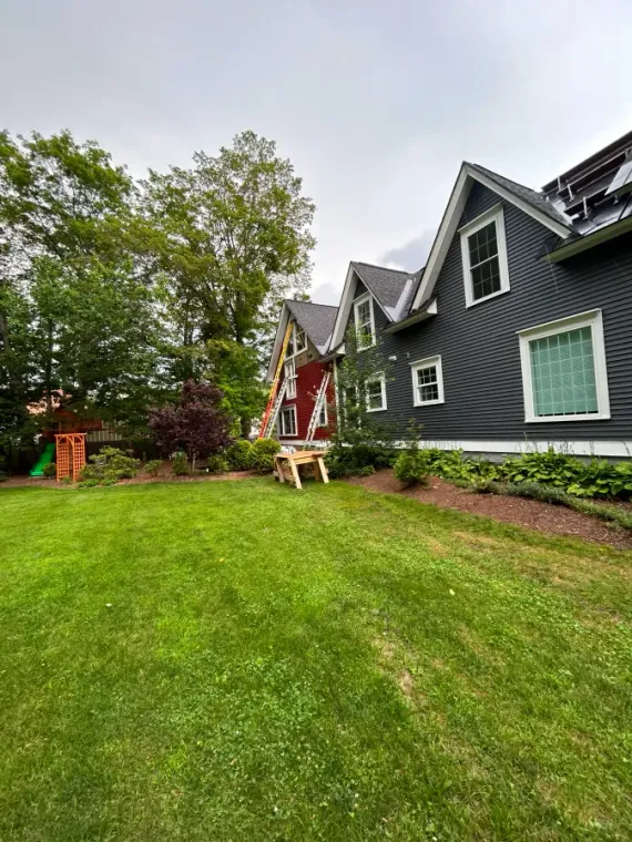 Green lawn beside a dark gray house with white trim and a small red porch in a wooded yard.