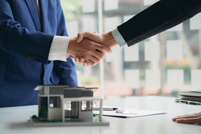 A man in a suit is shaking hands with another man in front of a model house.