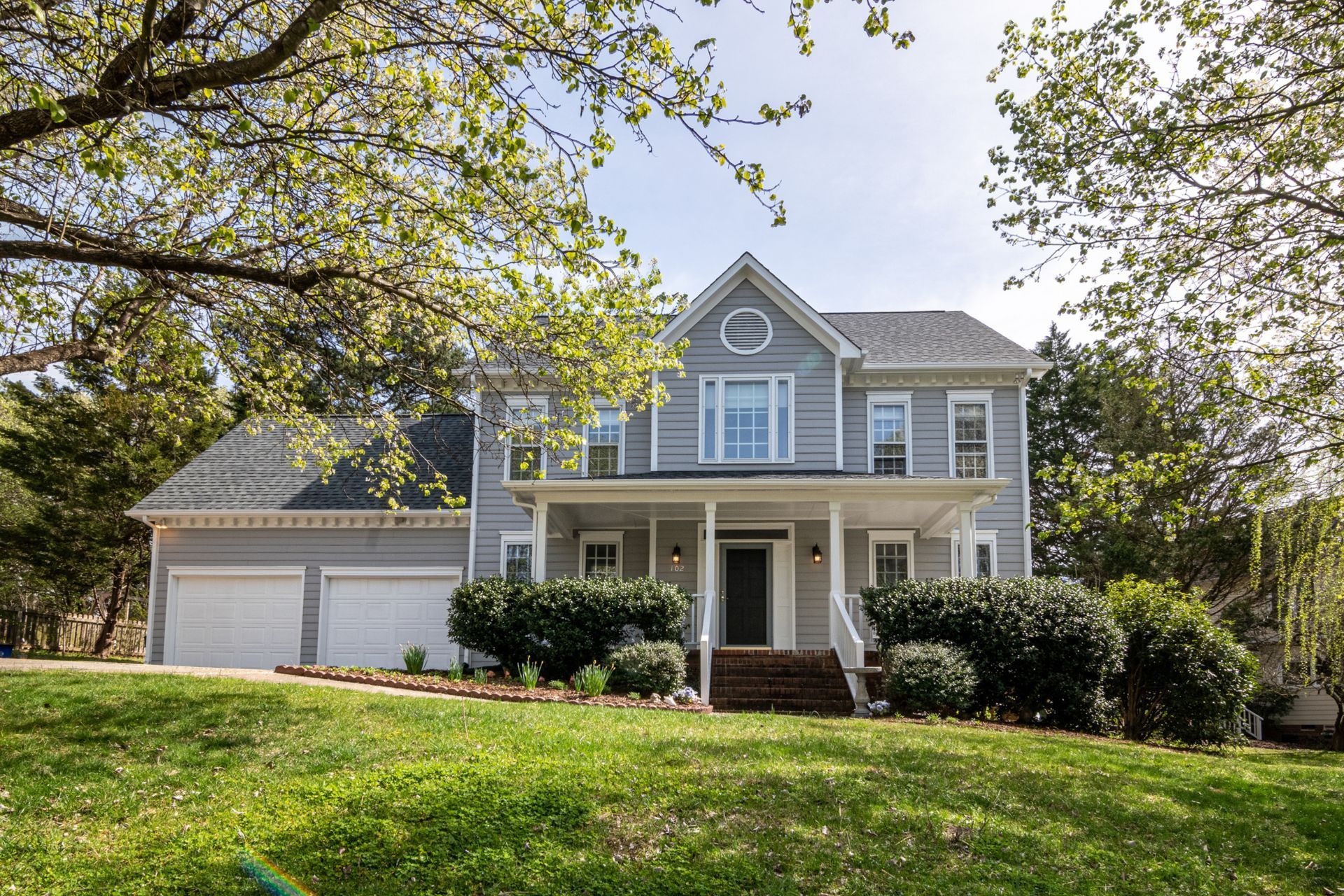 Gray two-story house with white trim, a front porch, and a two-car garage, on a green lawn with trees.