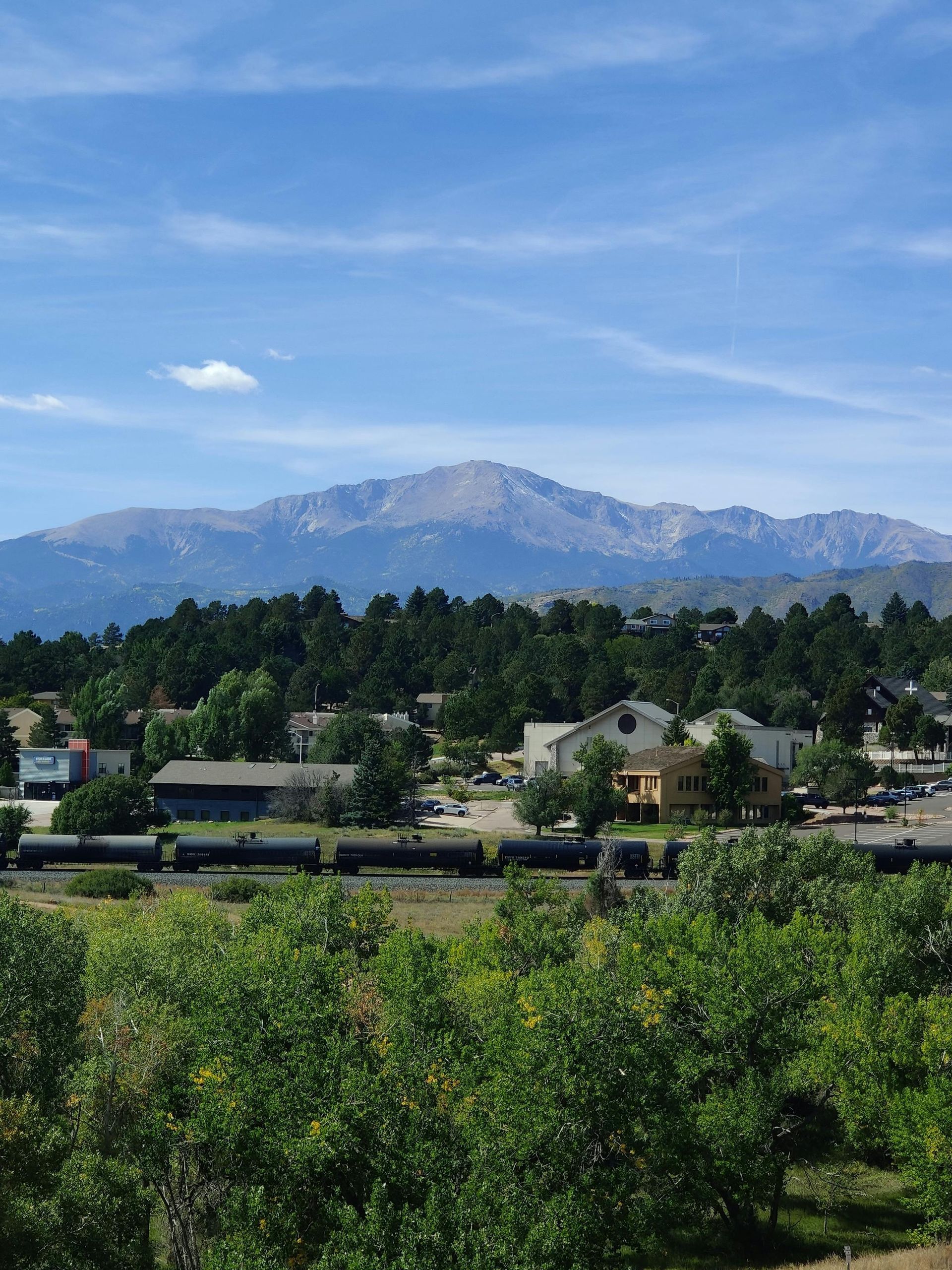 Town with train tracks, green trees, and a mountain under a blue sky.