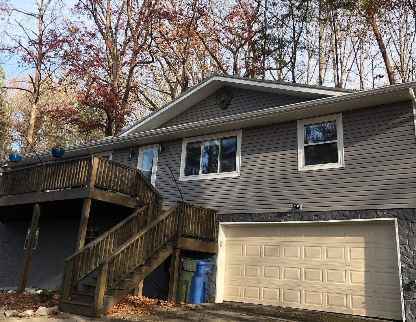 A gray split-level house with a tan garage door, a wooden deck, and stairs surrounded by trees in autumn.