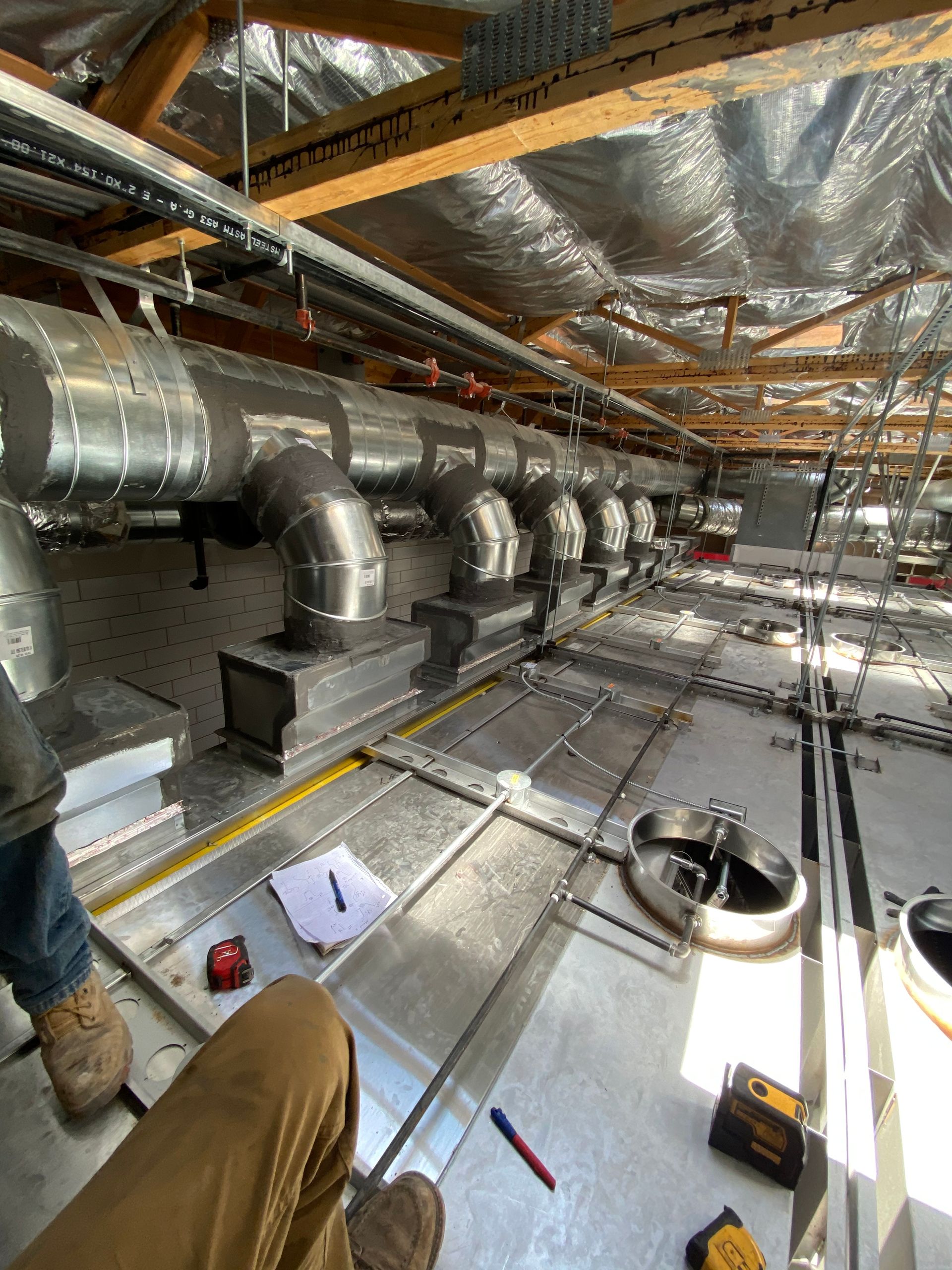 HVAC ductwork installation in a building's ceiling. Workers and tools visible. Silver ducts, metal ceiling.