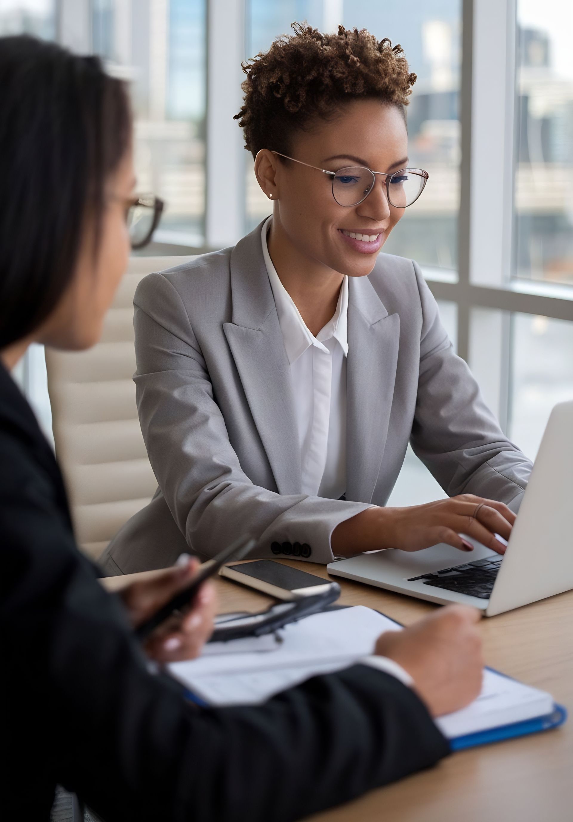 Professional woman in a grey blazer smiling while working on a laptop during a client consultation in a modern office.