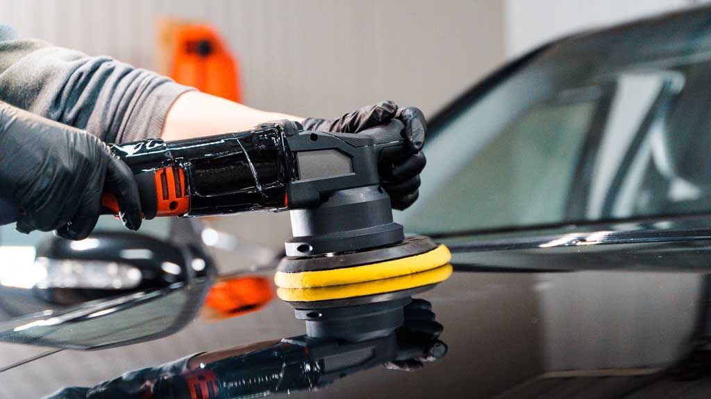 Person in black gloves using an electric polisher on a black car, polishing the surface.