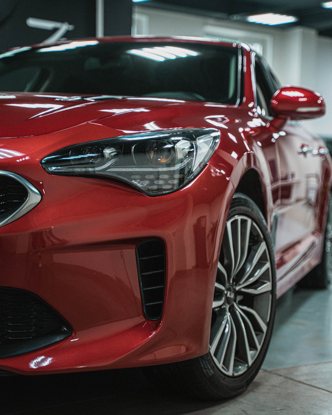 Red Kia Stinger car parked indoors, angled view of front, displaying its headlight and wheel.