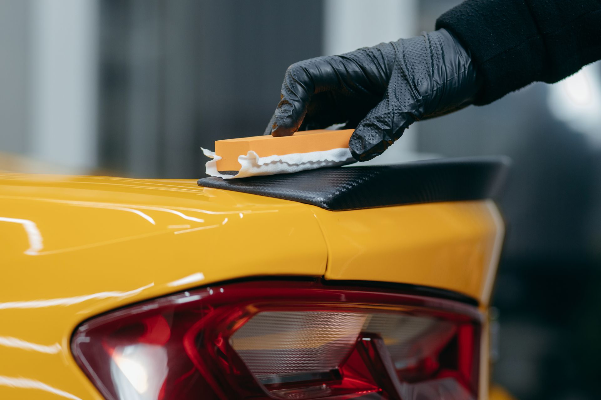 Gloved hand applying a coating with a sponge to a yellow car's spoiler.