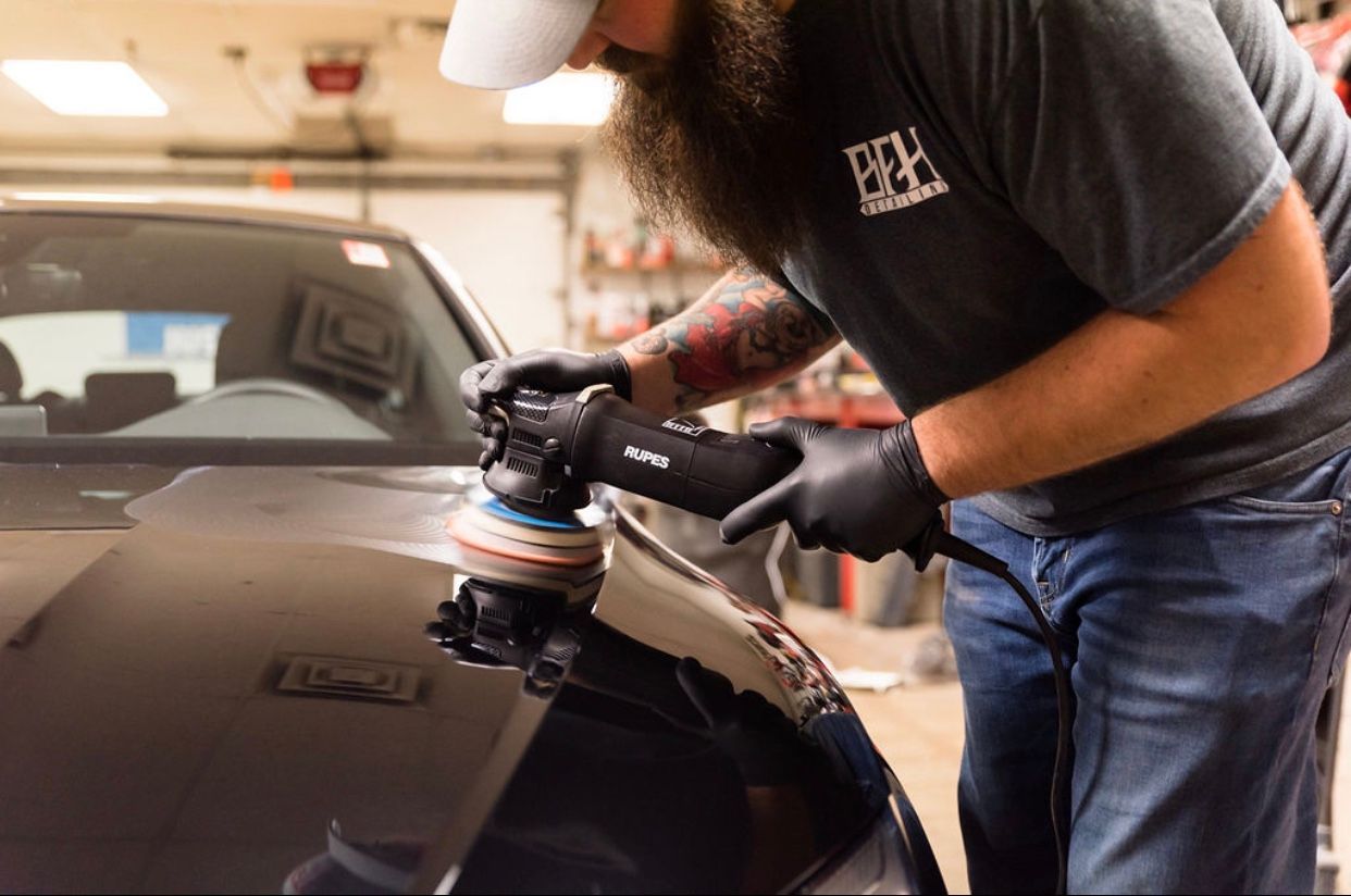 Man polishing a black car hood with an electric buffer in a garage.