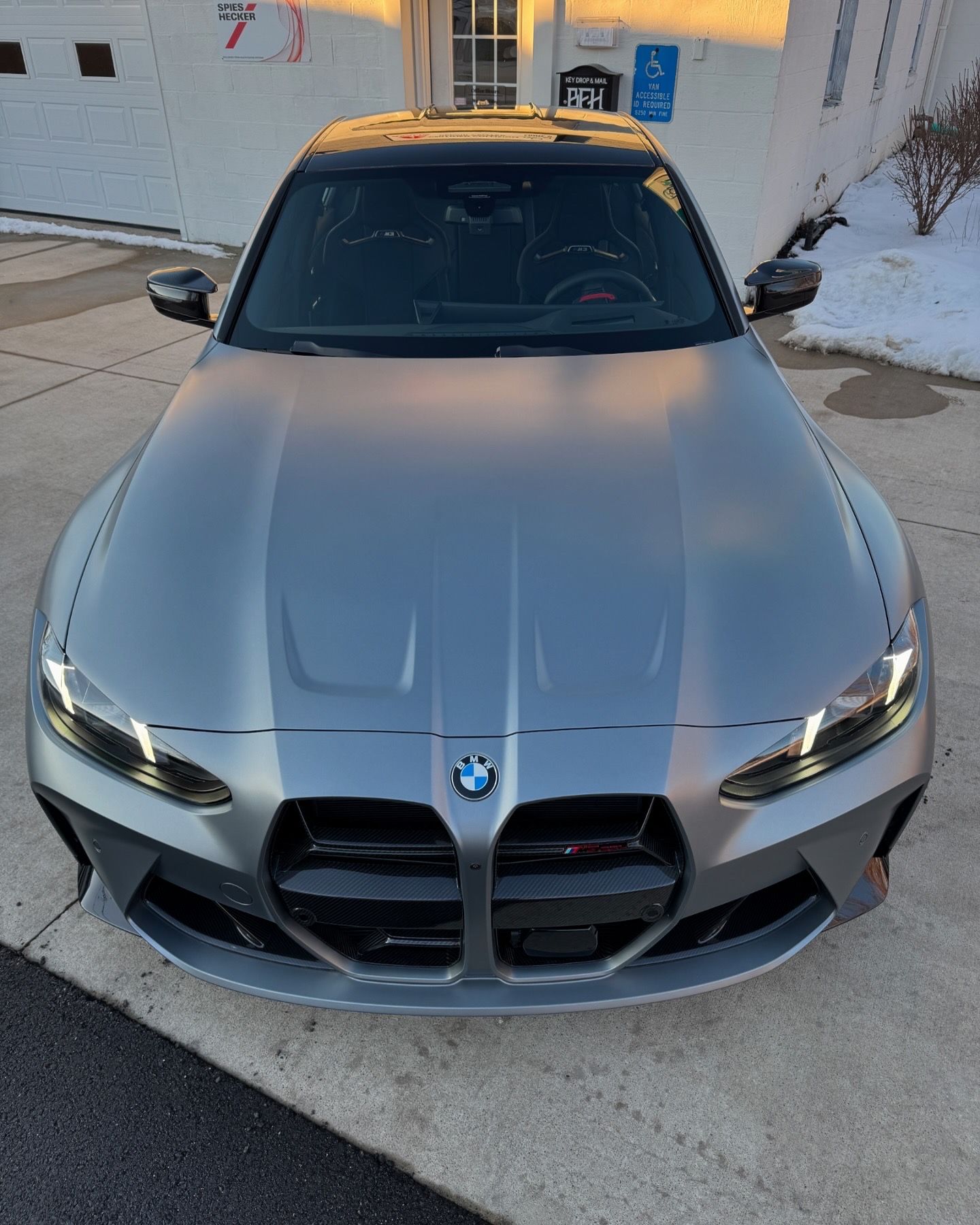 Gray BMW M3 sedan parked in front of a white building with snow.