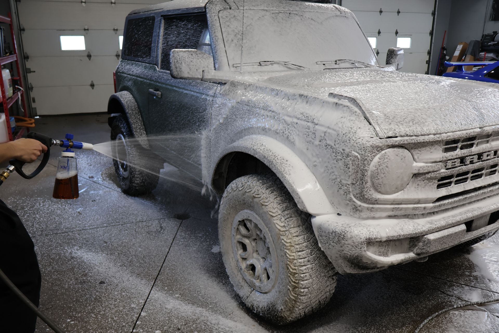 A person sprays soapy foam on a green Ford Bronco in a garage.