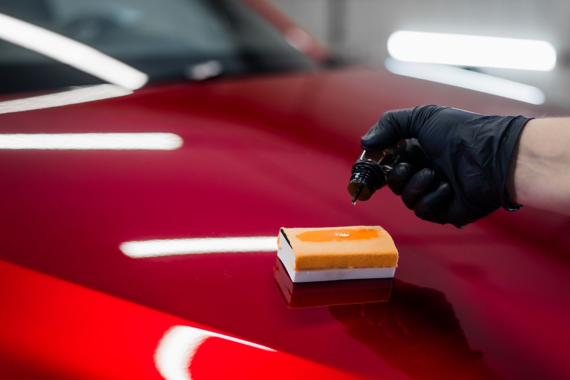 Hand in black glove applying ceramic coating to a red car hood with an applicator.
