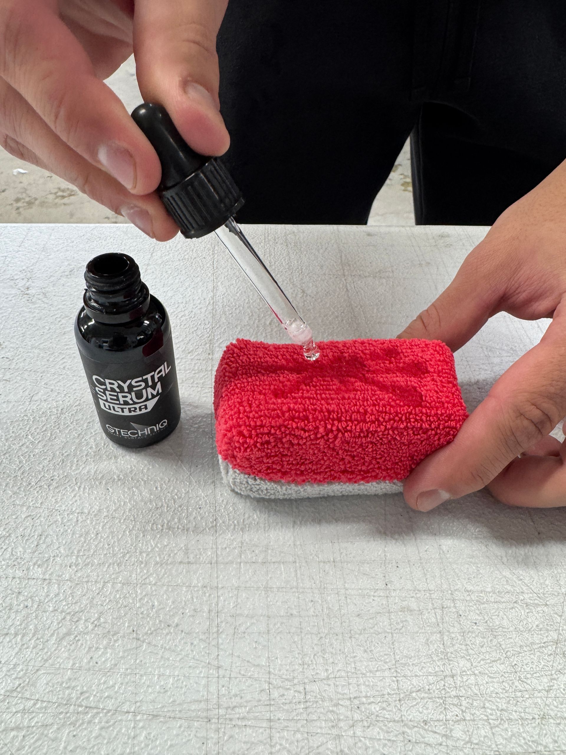 Person using a dropper to apply liquid from a black bottle onto a red microfiber pad on a gray surface.