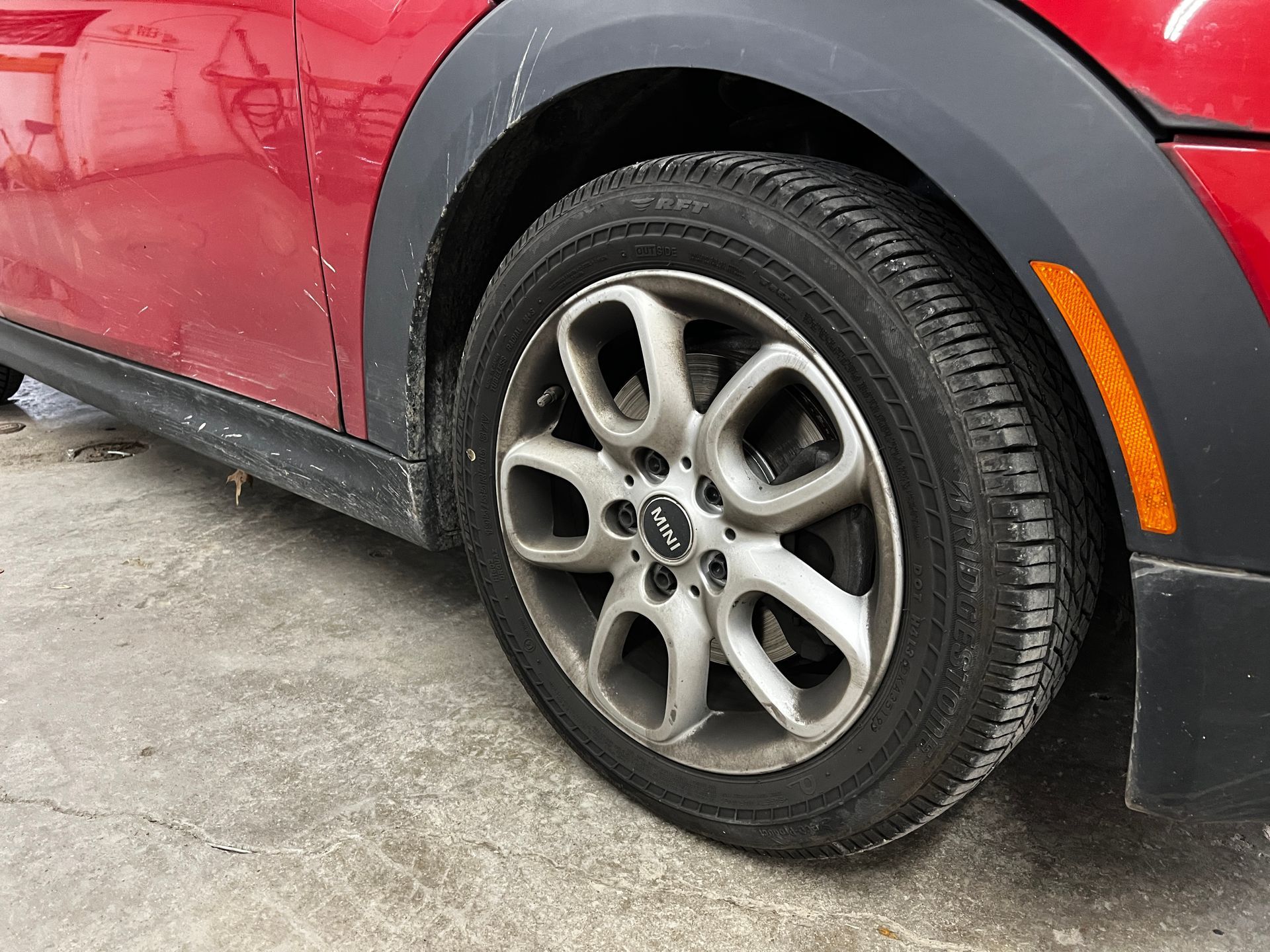 Red car's front tire, wheel, and fender. Tire is dirty; wheel is silver. Amber side marker light visible.