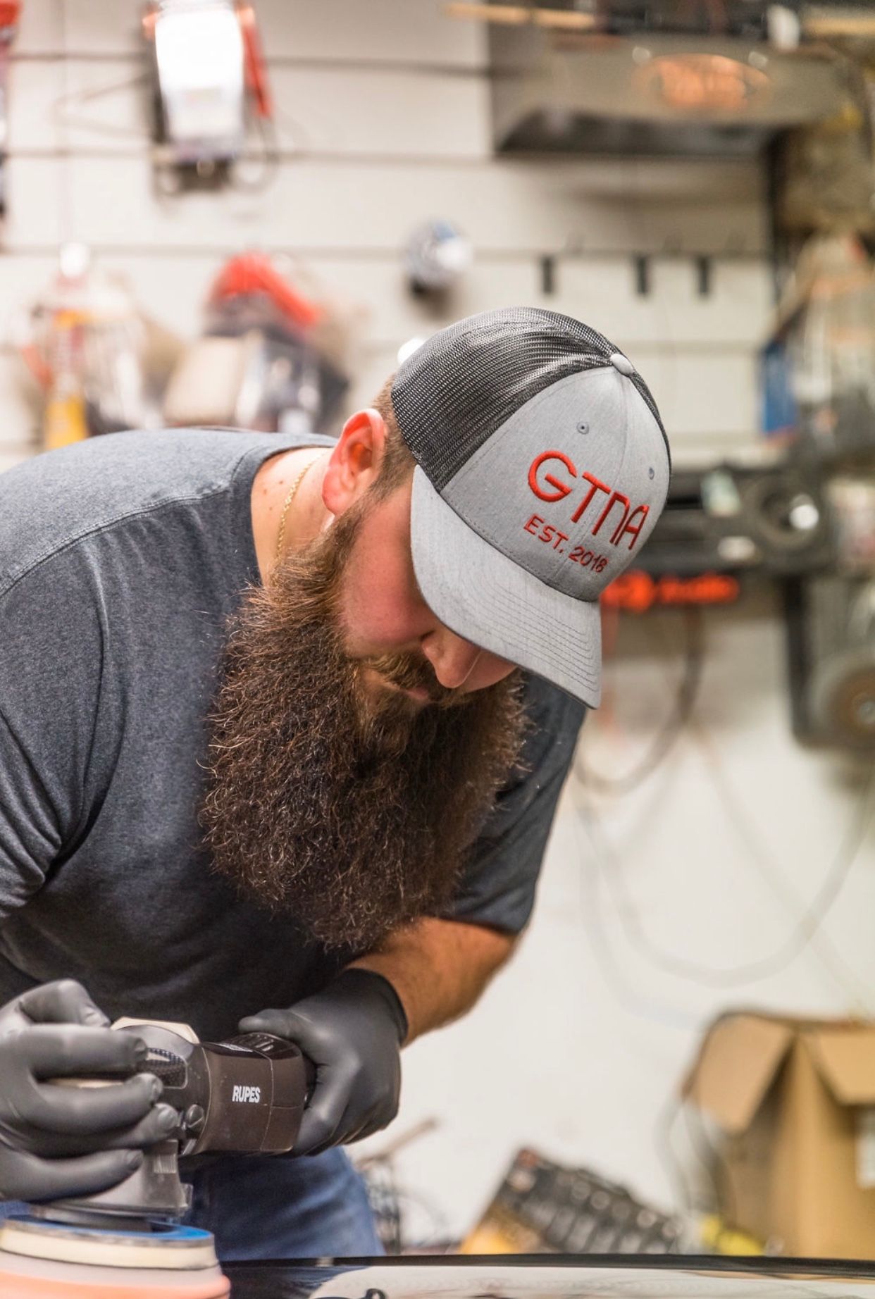 Man with a long beard wearing a hat and gloves polishing a dark surface in a workshop.